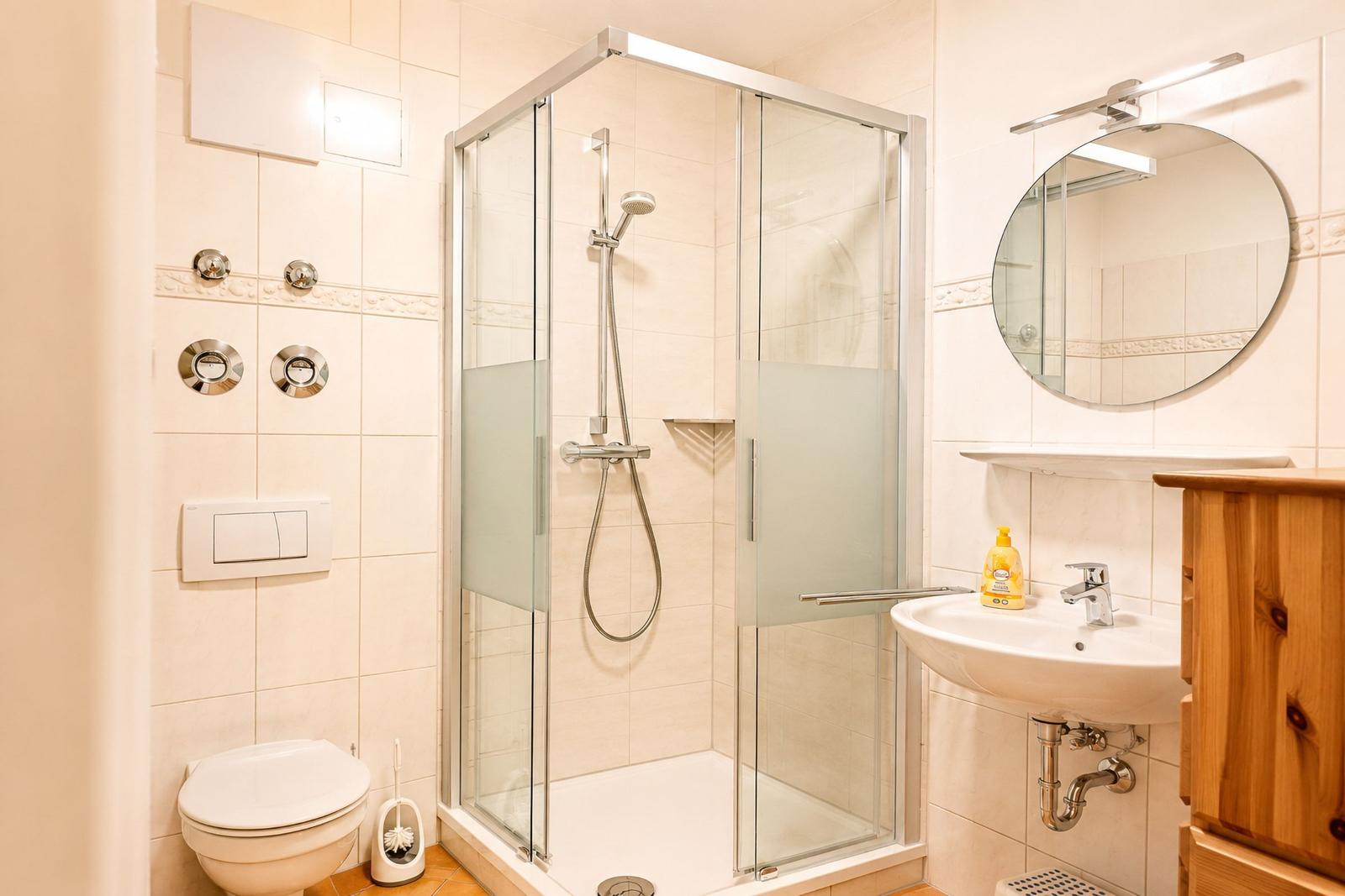 Badezimmer mit Dusche, Waschbecken und Toilette. Holzschrank und runder Spiegel.