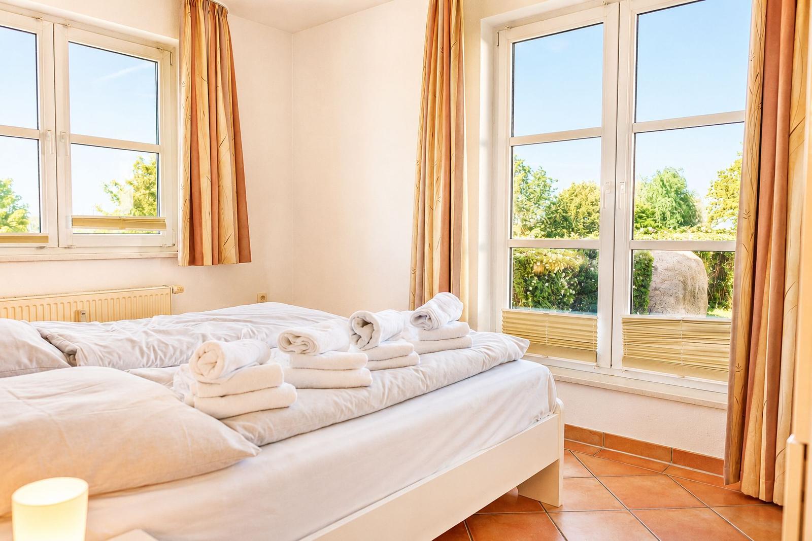 Schlafzimmer mit Bett, Handtüchern und Fenstern mit Blick auf Garten.