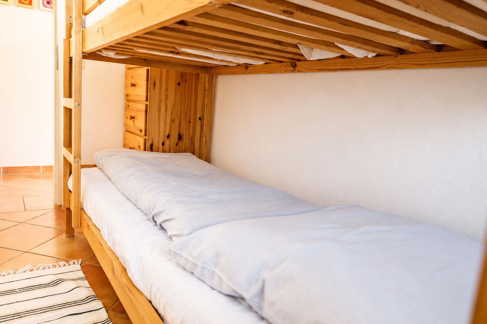 Unterstockbett mit Matratze und Bettwäsche. Holzgestell und Nachttisch sichtbar.