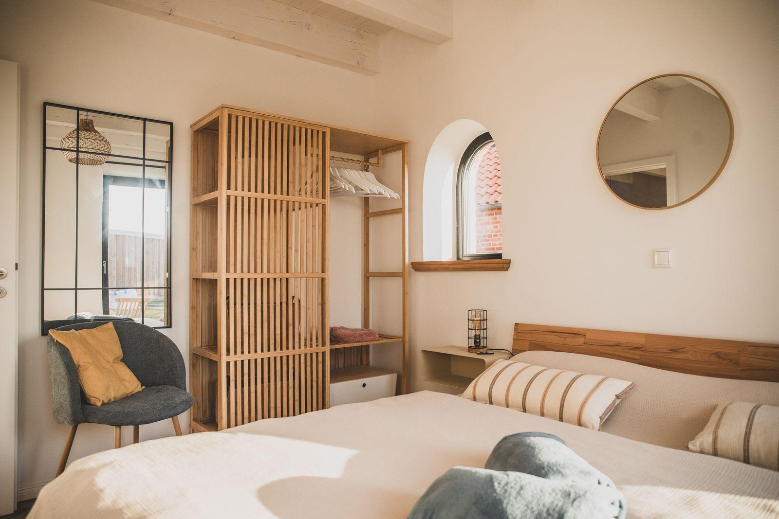 Schlafzimmer mit Bett, Holzgarderobe, runden Spiegel und Bogenfenster.