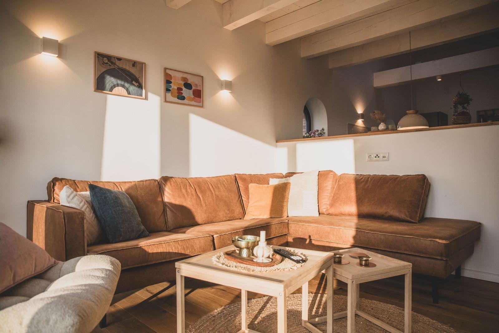Ein Wohnzimmer mit braunem Sofa, Holztischen und Wandbildern. Holzbalken und warme Beleuchtung.