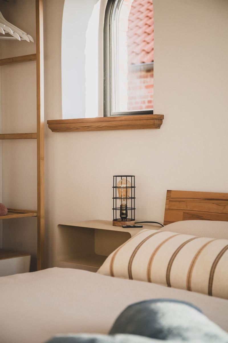 Schlafzimmer mit Bett, Nachttisch und Fenster. Holzregal und Lampe sind sichtbar.