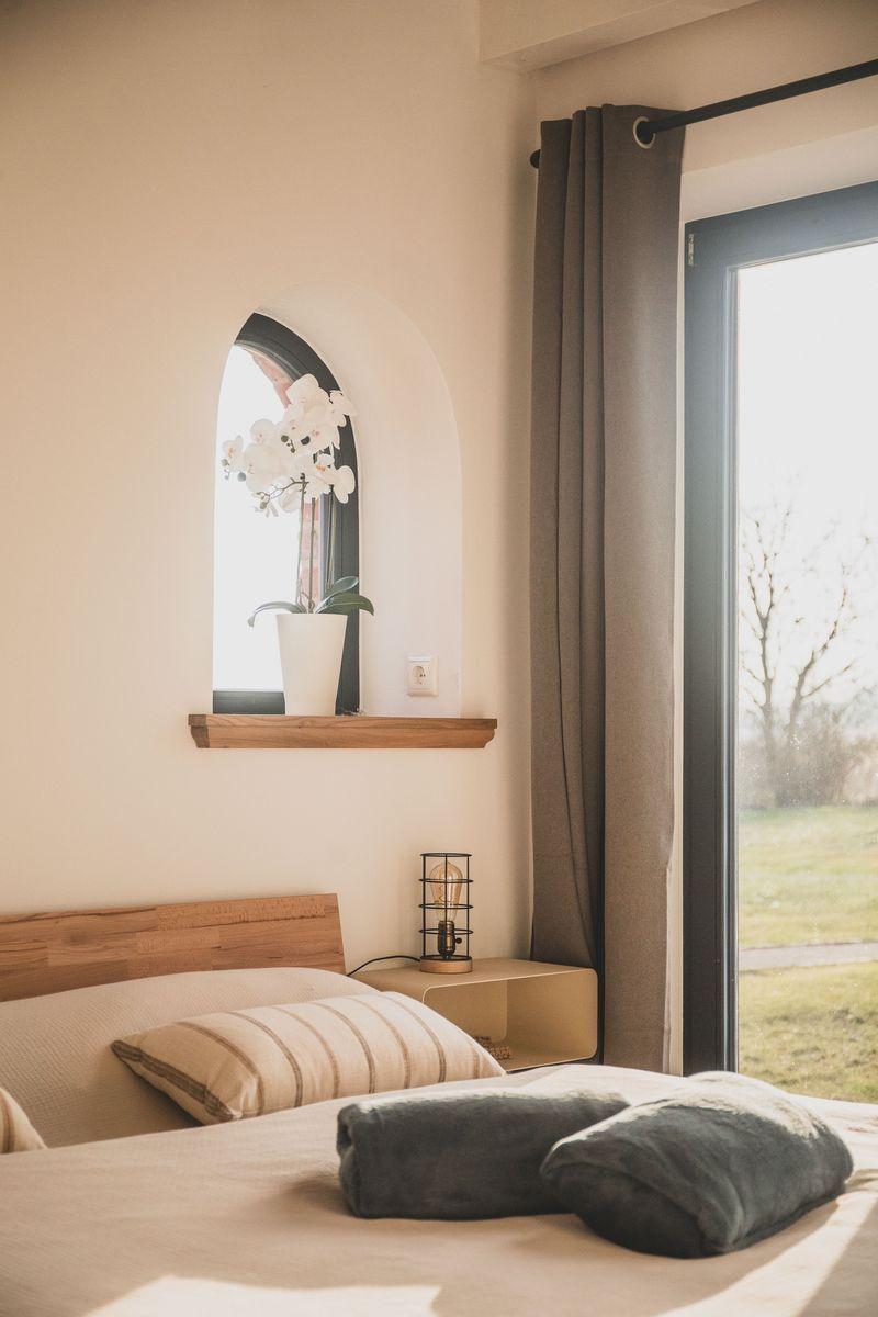 Schlafzimmer mit Bett, Nachttisch und Fenster. An der Wand hängt ein Spiegel mit Orchidee.