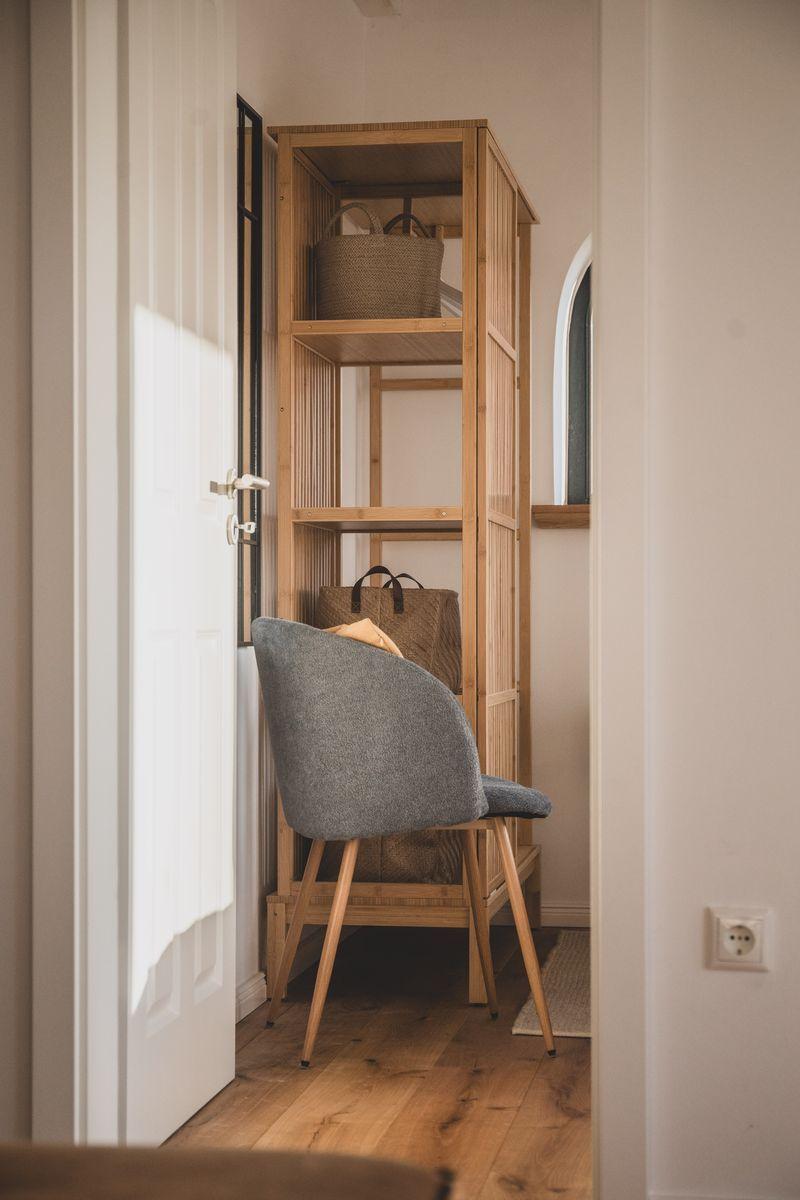 Ein grauer Stuhl steht vor einem hohen Holzregal mit Körben. Eine Tür und ein Fenster sind sichtbar.