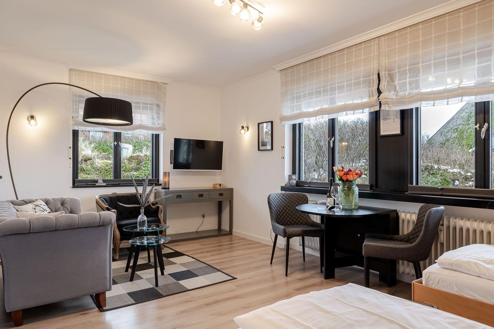 Living space with sofa, TV, dining table, and bed. Windows with blinds and view of garden.