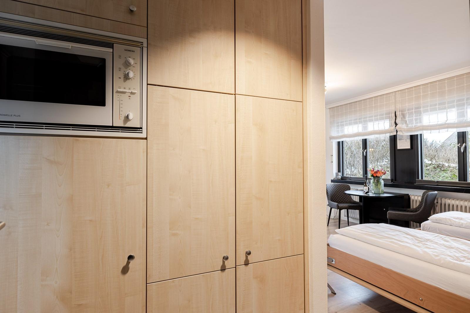Kitchen with integrated microwave and wooden cabinets. In the background, a bedroom with a bed and table.