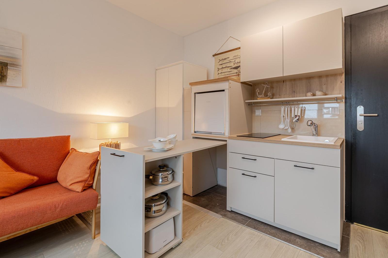 Küche mit Arbeitsfläche, Spüle und Kühlung. Nebenraum mit Sofa und Lampe.