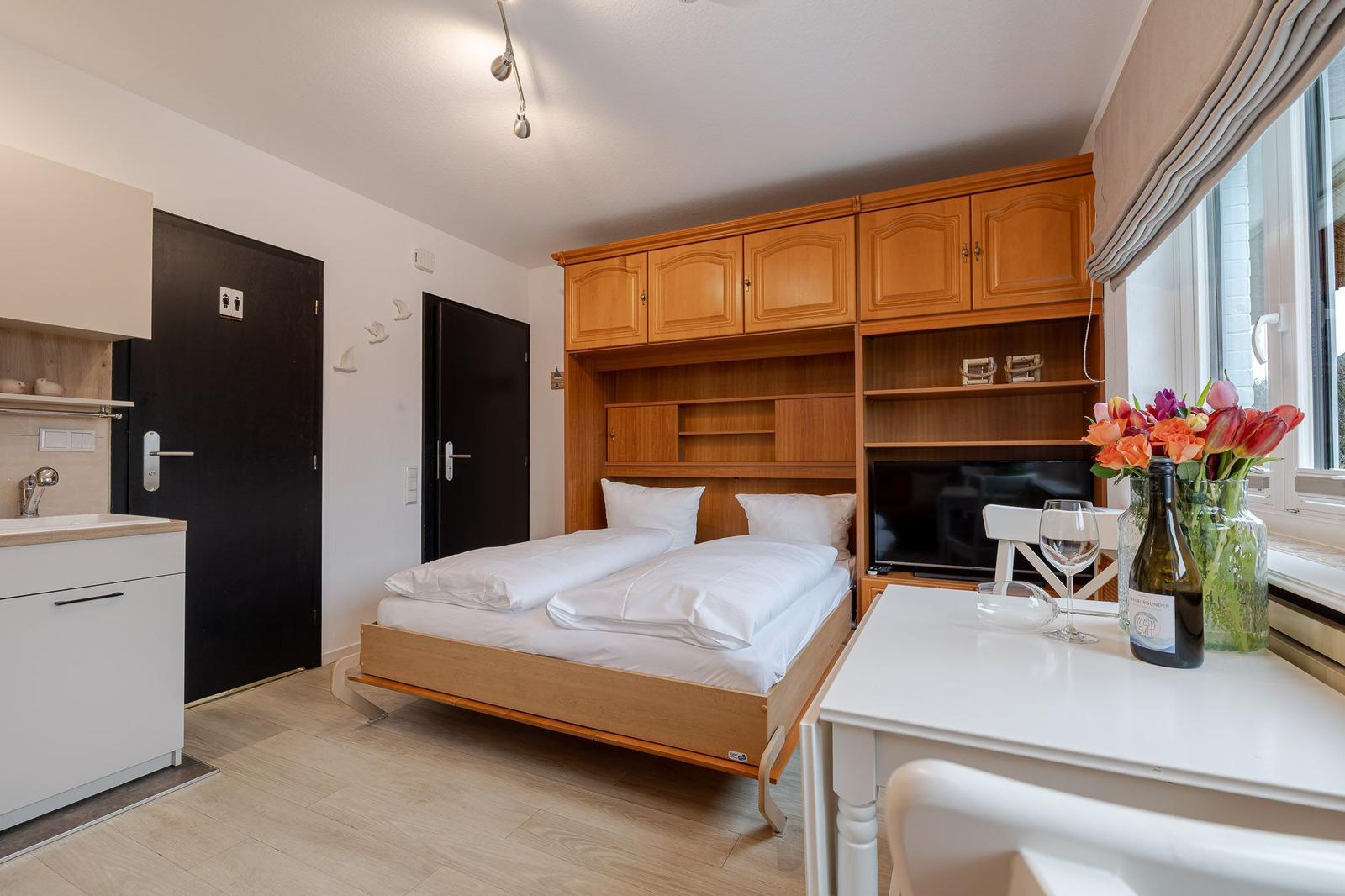 Schlafzimmer mit Bett, Küchenzeile und TV. Fenster mit Blumen und Weinflasche.