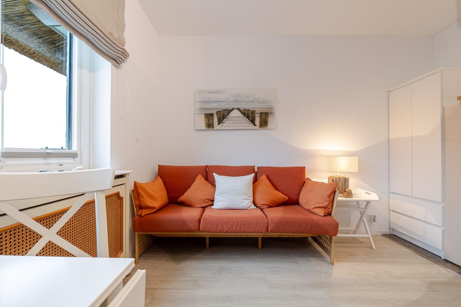 Ein Raum mit orangefarbenem Sofa, weißen Möbeln und einem Fenster mit Jalousie.