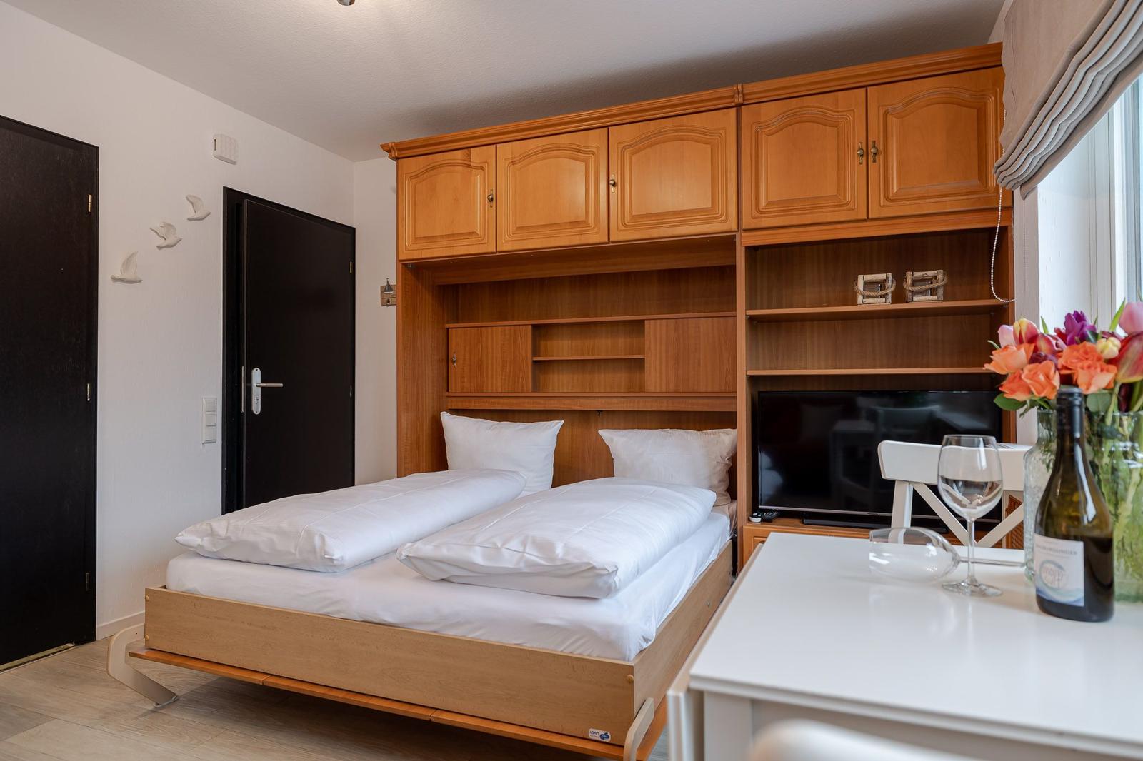 Schlafzimmer mit Bett, Schränken und Fernseher. Tisch mit Wein und Blumen.