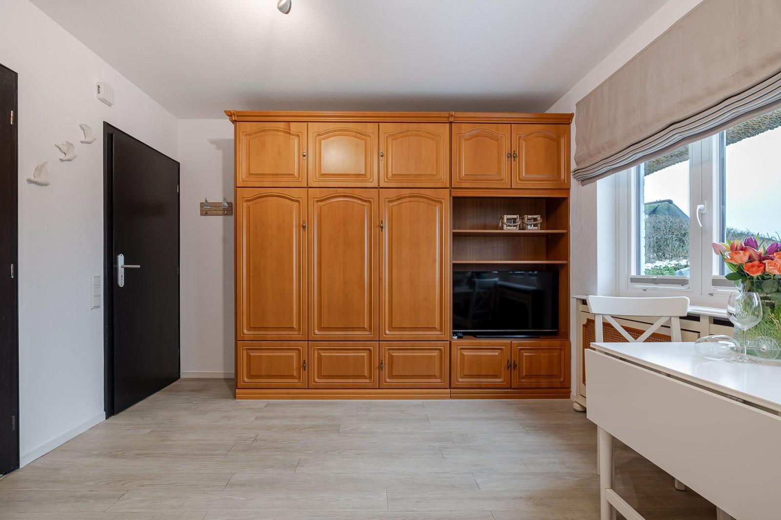 Großer Schrank mit TV-Nische, Fenster mit Blumen, weiße Tischplatte und Holzboden.