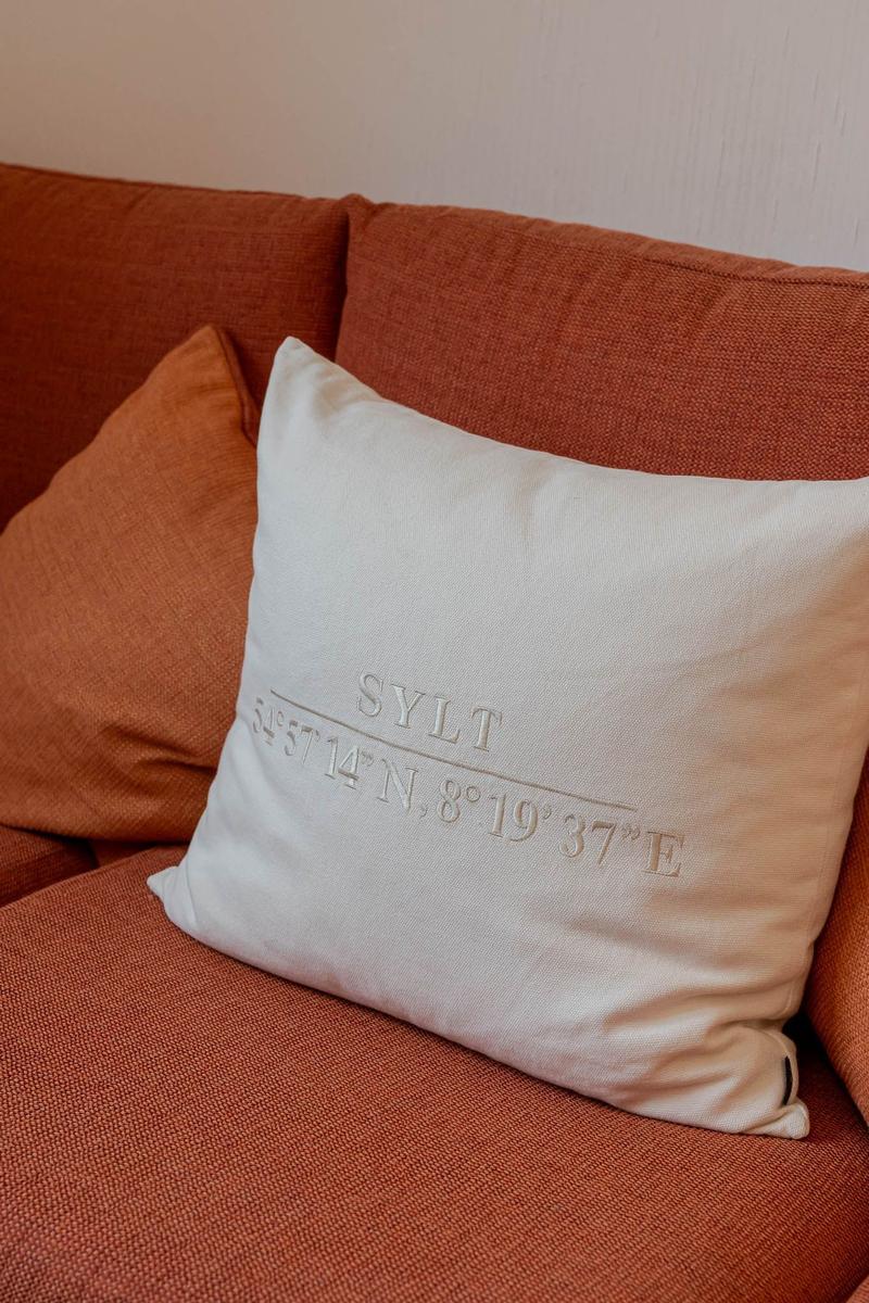 Weißer Kissenbezug mit geprägtem Ortsnamen und Koordinaten auf einem orangefarbenen Sofa.