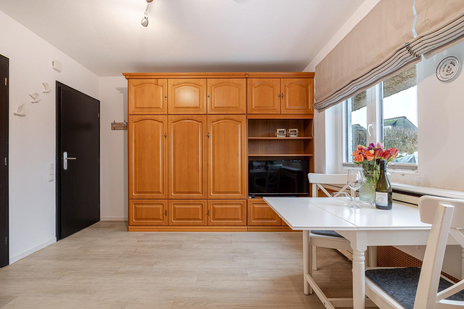 Wohn- und Essbereich mit Holzschrank, TV und weißem Tisch mit Stühlen.