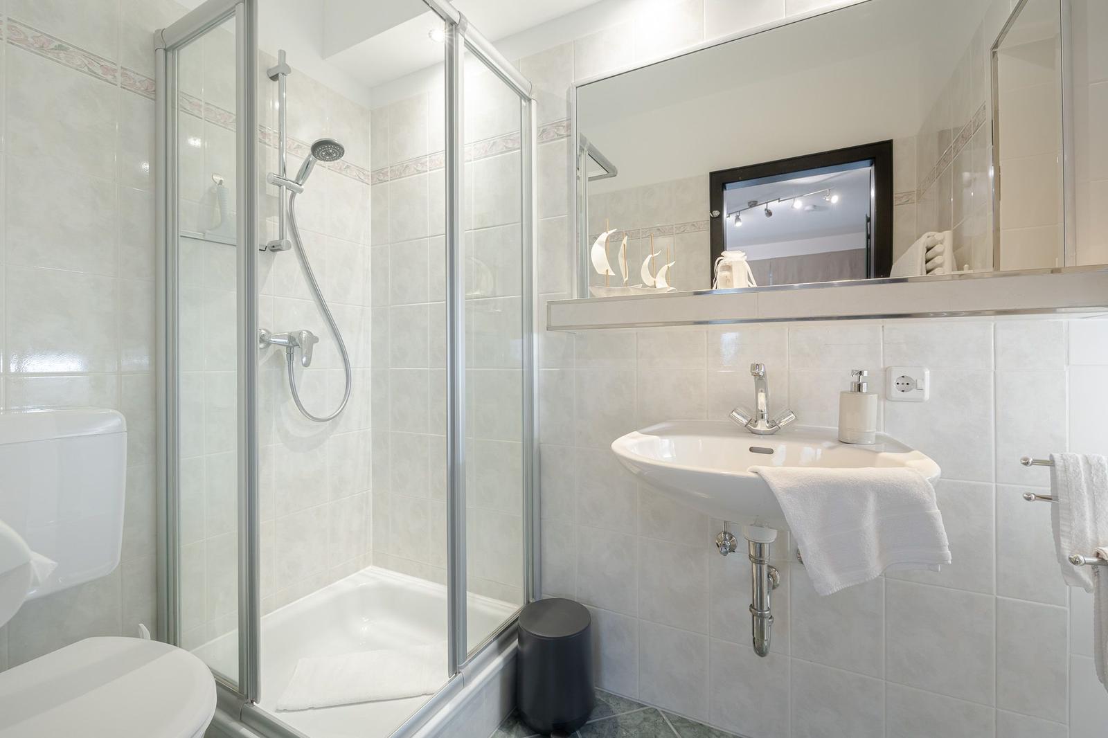 Badezimmer mit Dusche, Waschbecken und Spiegel. Weiße Fliesen und Handtuchhalter.
