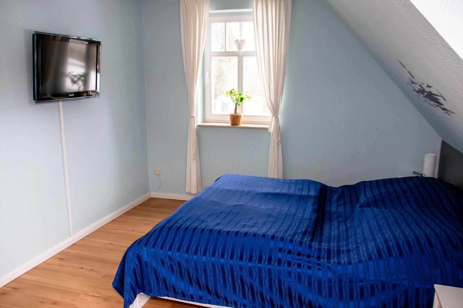 Schlafzimmer mit blauem Bett, Wandfernseher und Fenster mit Vorhängen.