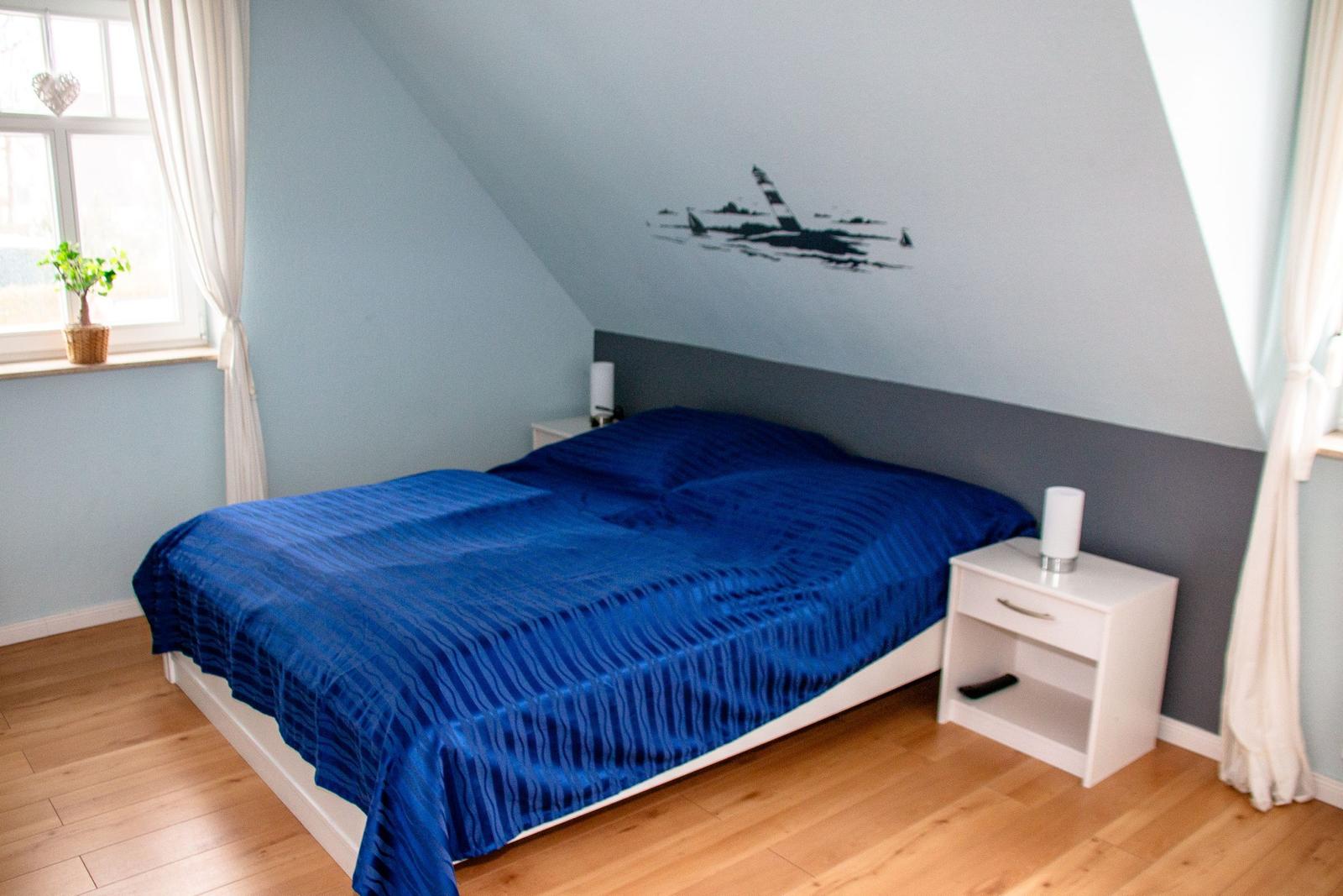 Schlafzimmer mit Doppelbett, blauer Bettwäsche, Holzboden und Fenster mit Vorhang.