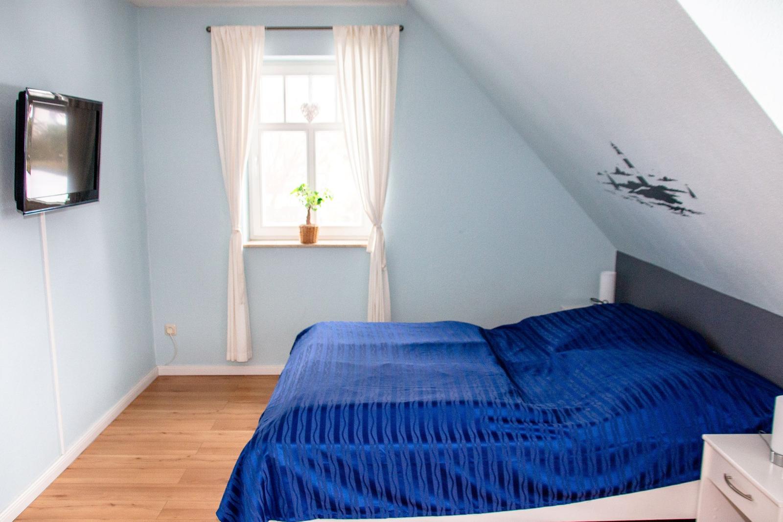 Schlafzimmer mit Dachschräge, blauem Bett und Fenster mit weißen Vorhängen.