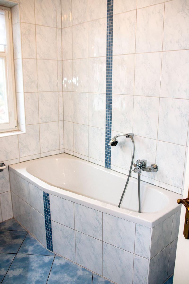 Bad mit Badewanne, Duschkopf und Fenster. Fliesen mit blauer Streifenverzierung.