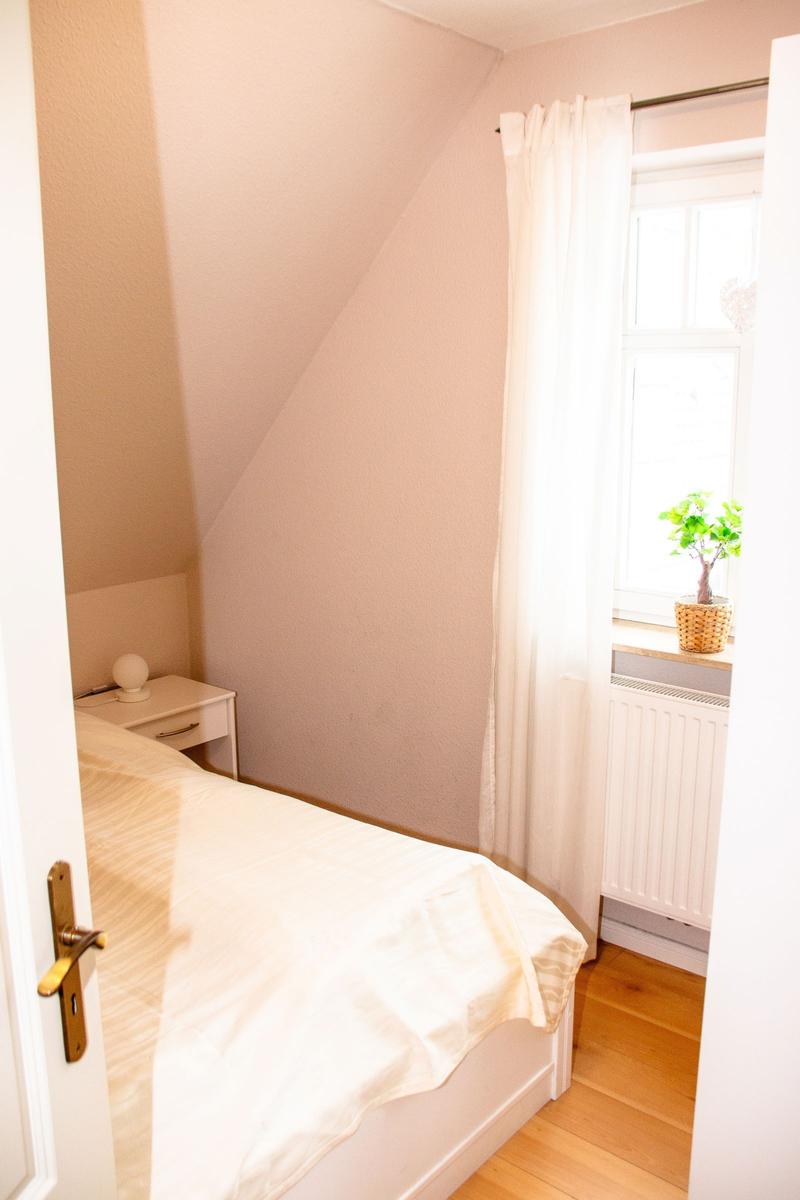 Schlafzimmer mit Bett, Nachttisch und Fenster mit Vorhang und Pflanze.