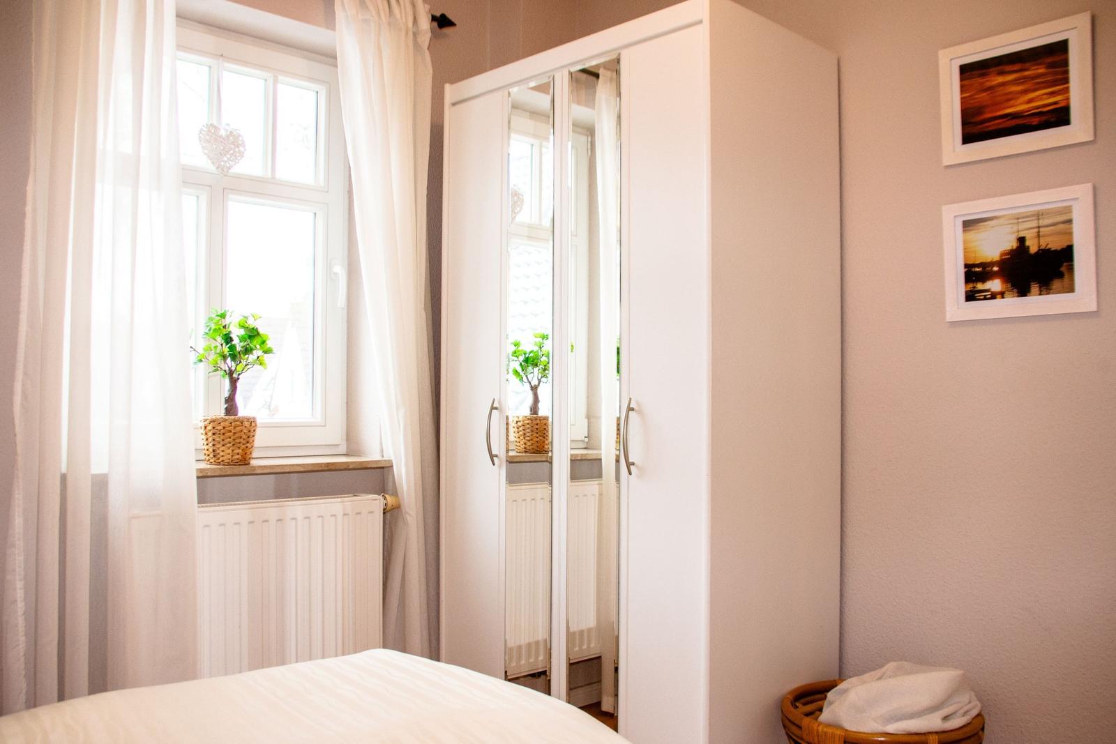 Schlafzimmer mit weißem Kleiderschrank, Fenster mit Vorhängen und Pflanze auf dem Fensterbrett.