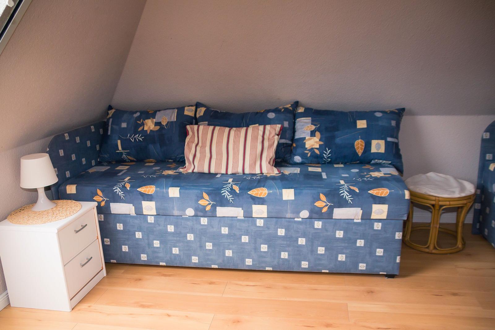 Schlafsofa mit blauem Stoff und Kissen, weiße Nachttischlampe, Holzboden und schräger Wand.