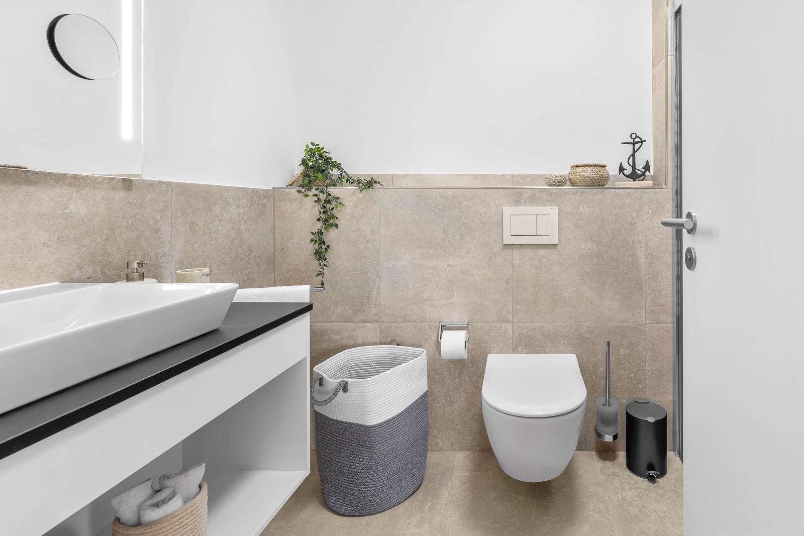 Badezimmer mit Waschbecken, Toilette und Dusche. Wandverkleidung und Bodenfliesen in Beige.