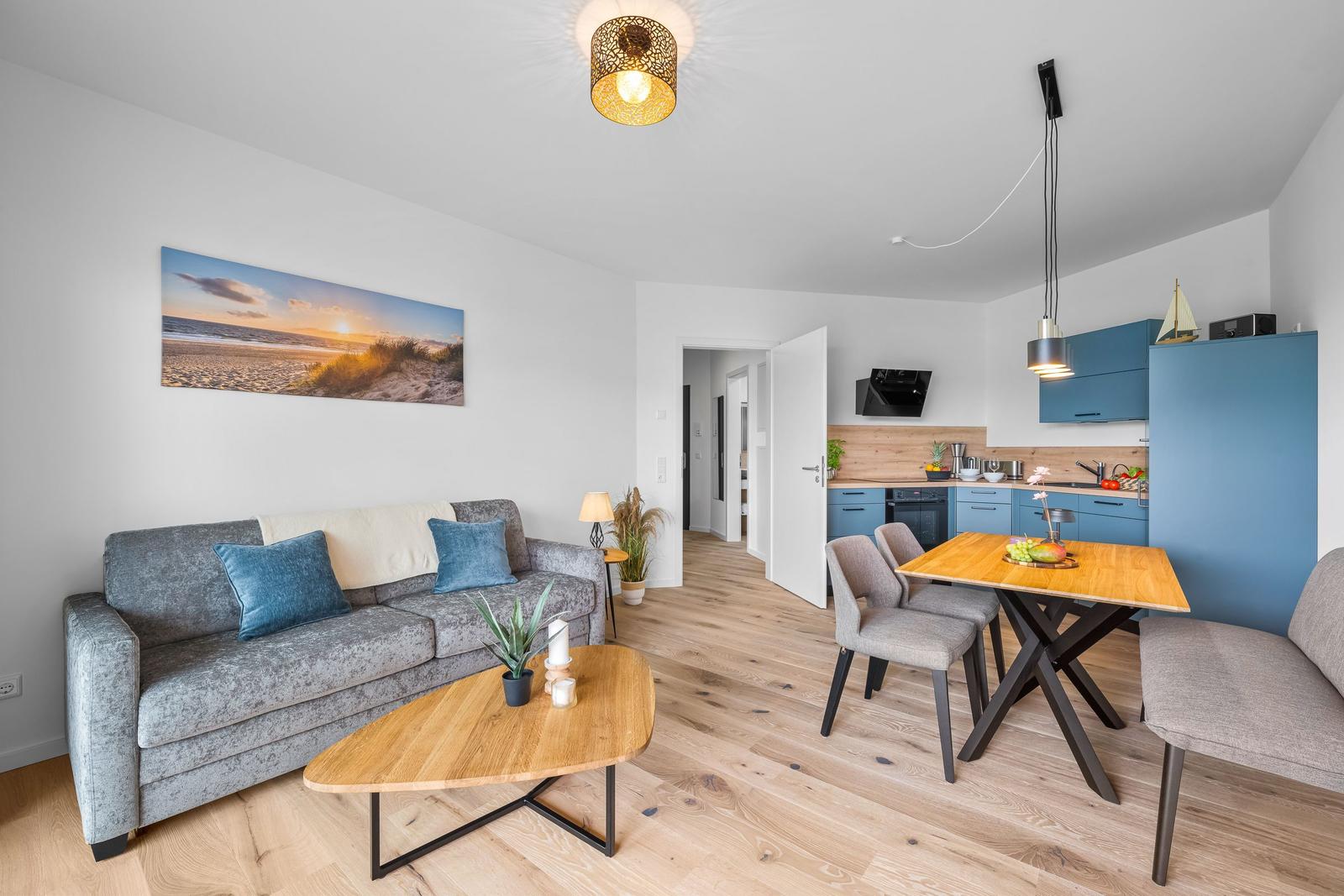 Wohnbereich mit Sofa, Esstisch und Küchenzeile. Holzboden und blaue Schränke.