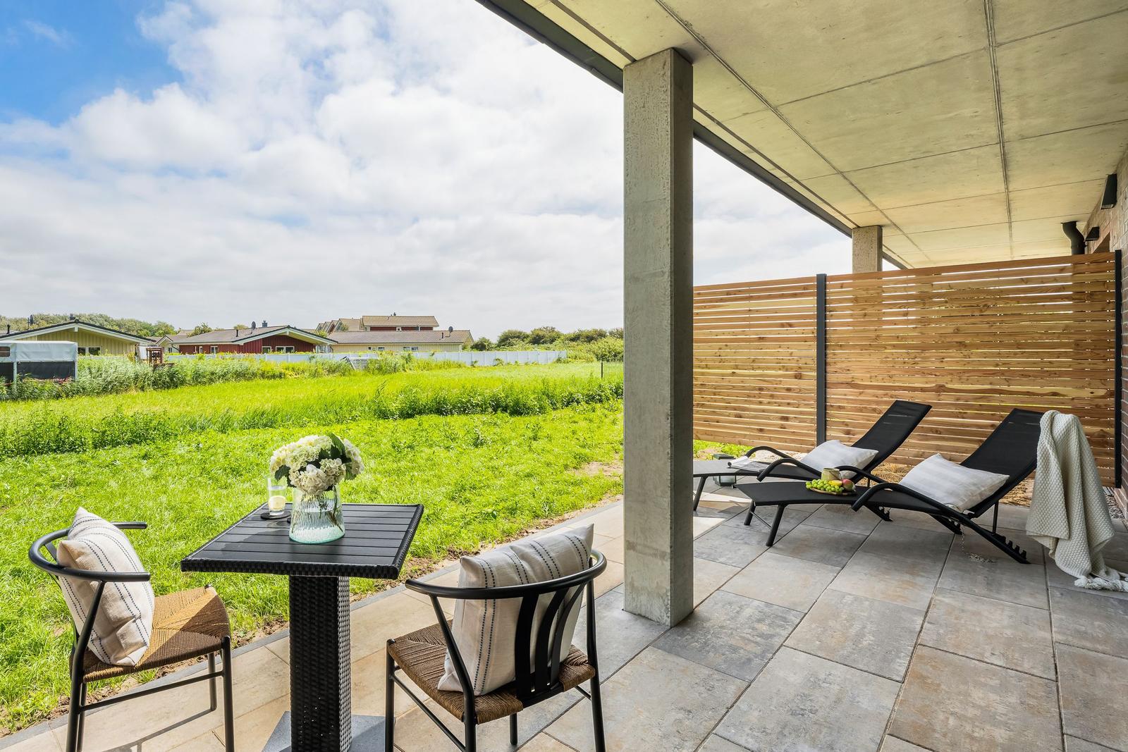 Terrasse mit Tisch, Stühlen und Liegen. Blick auf grünes Feld und Himmel.