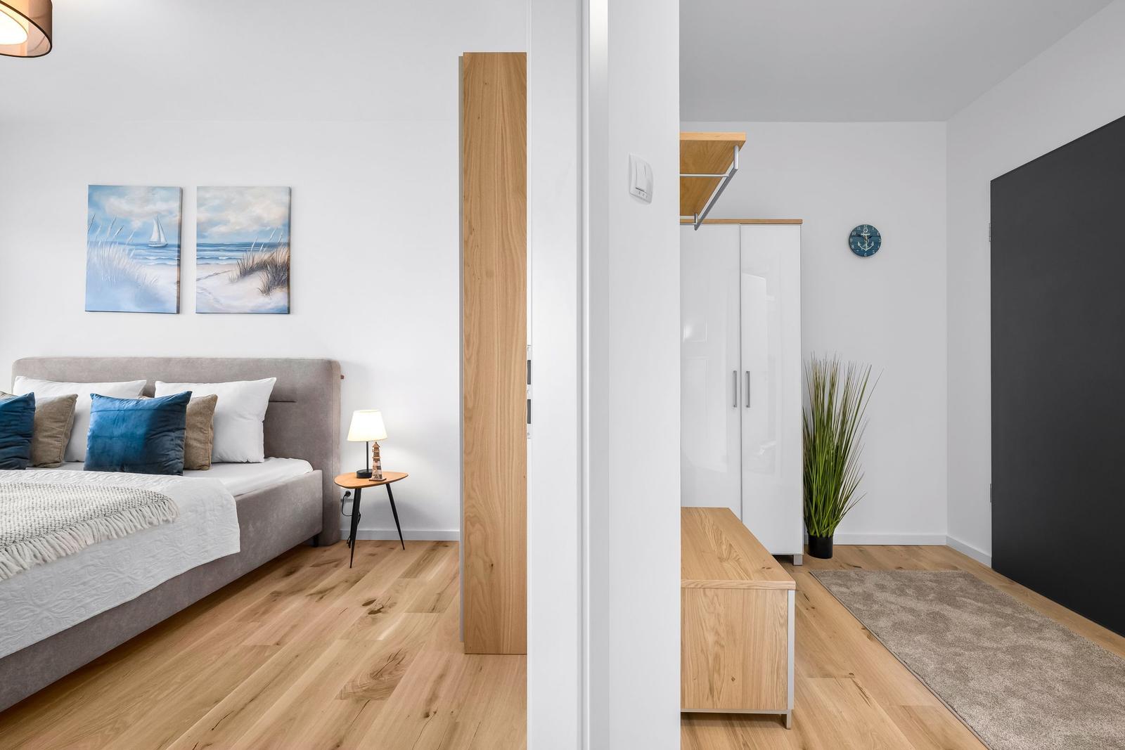 Schlafzimmer mit Bett, Wandbildern und Holzboden. Flur mit Schrank, Pflanze und Teppich.