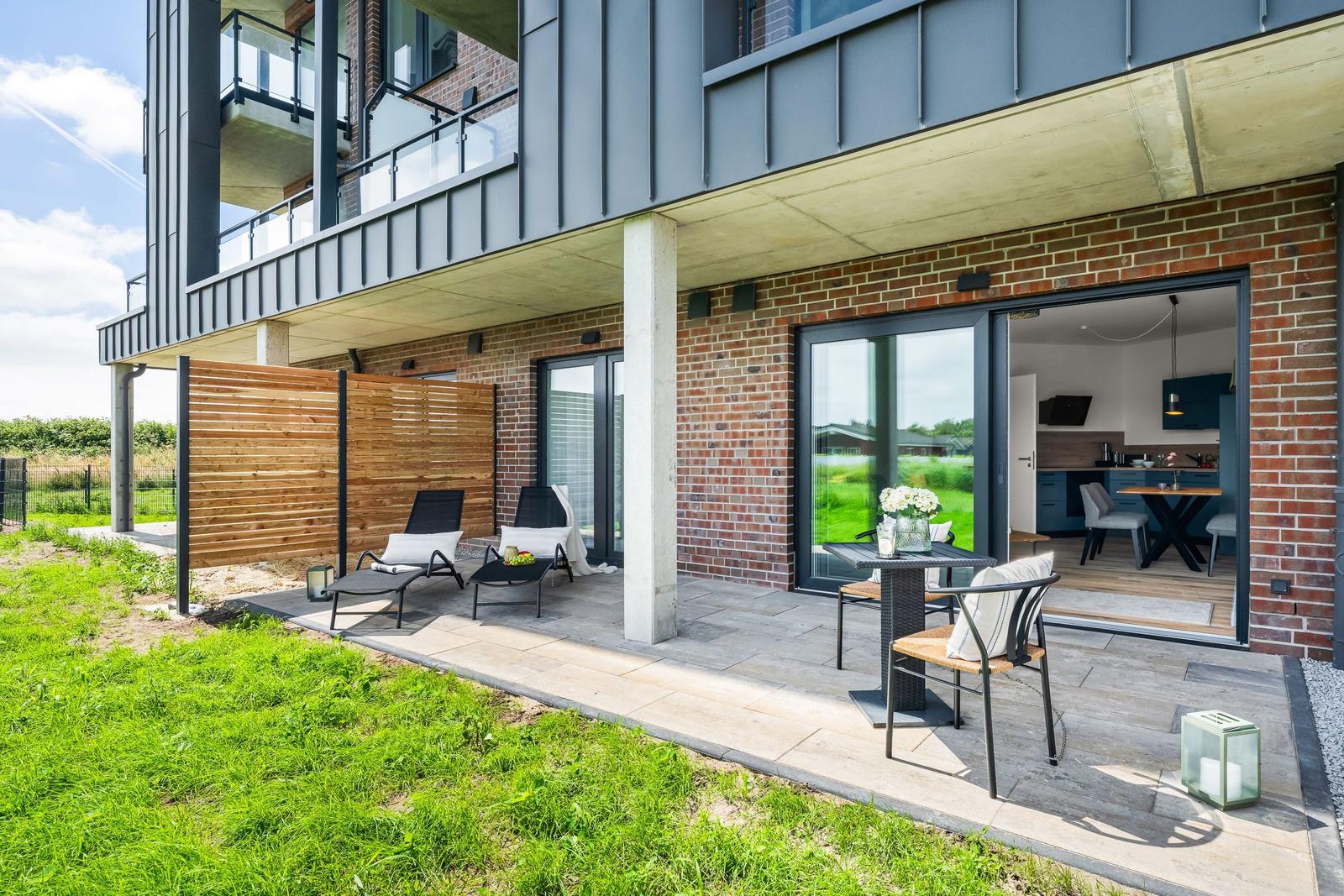 Terrasse mit Liegen und Tisch. Durchgehende Glastüren führen ins Haus. Hintergrund: grünes Feld.