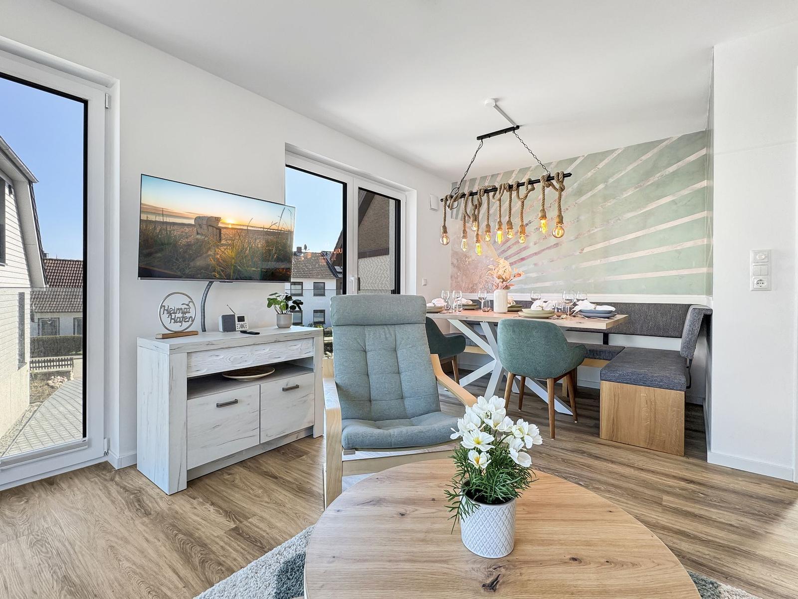 Wohn- und Essbereich mit TV, Sitzgelegenheiten und Essplatz. Holzboden und große Fenster.