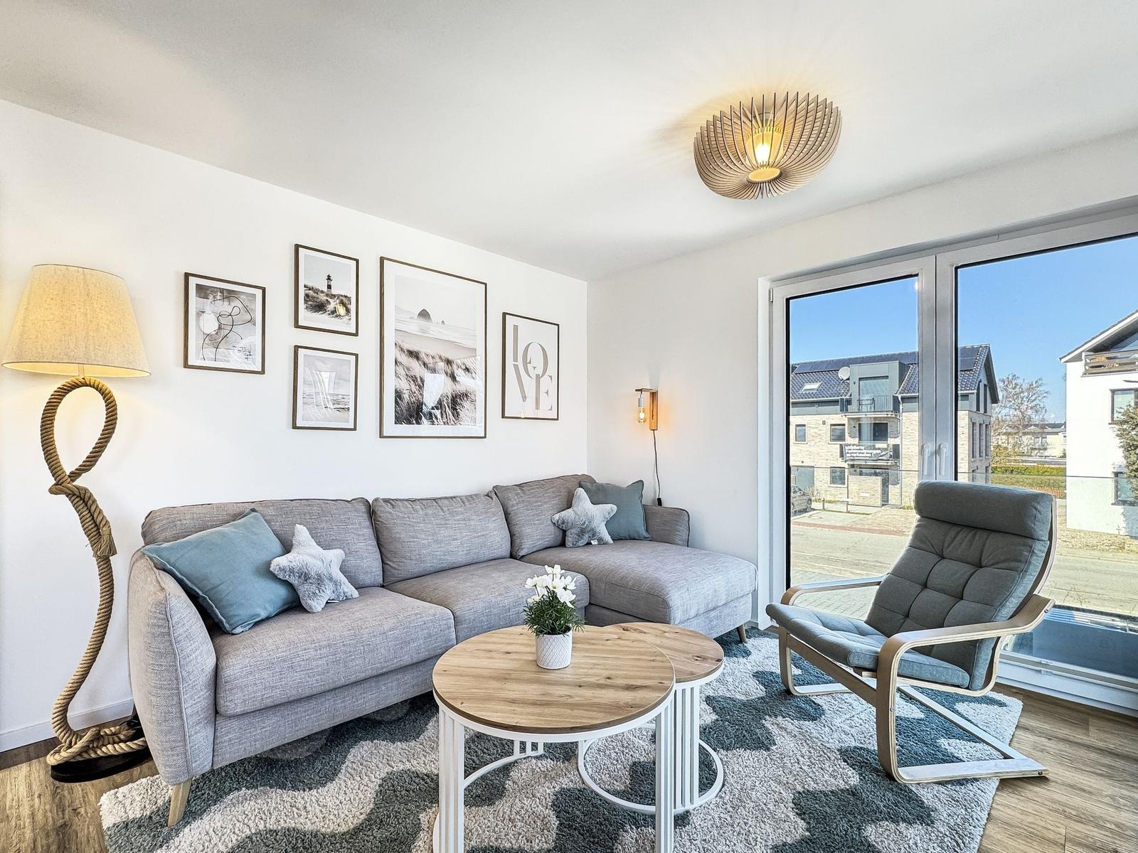 Wohnzimmer mit Sofa, Sessel, Tisch und Wandbildern. Große Fenster mit Blick nach draußen.