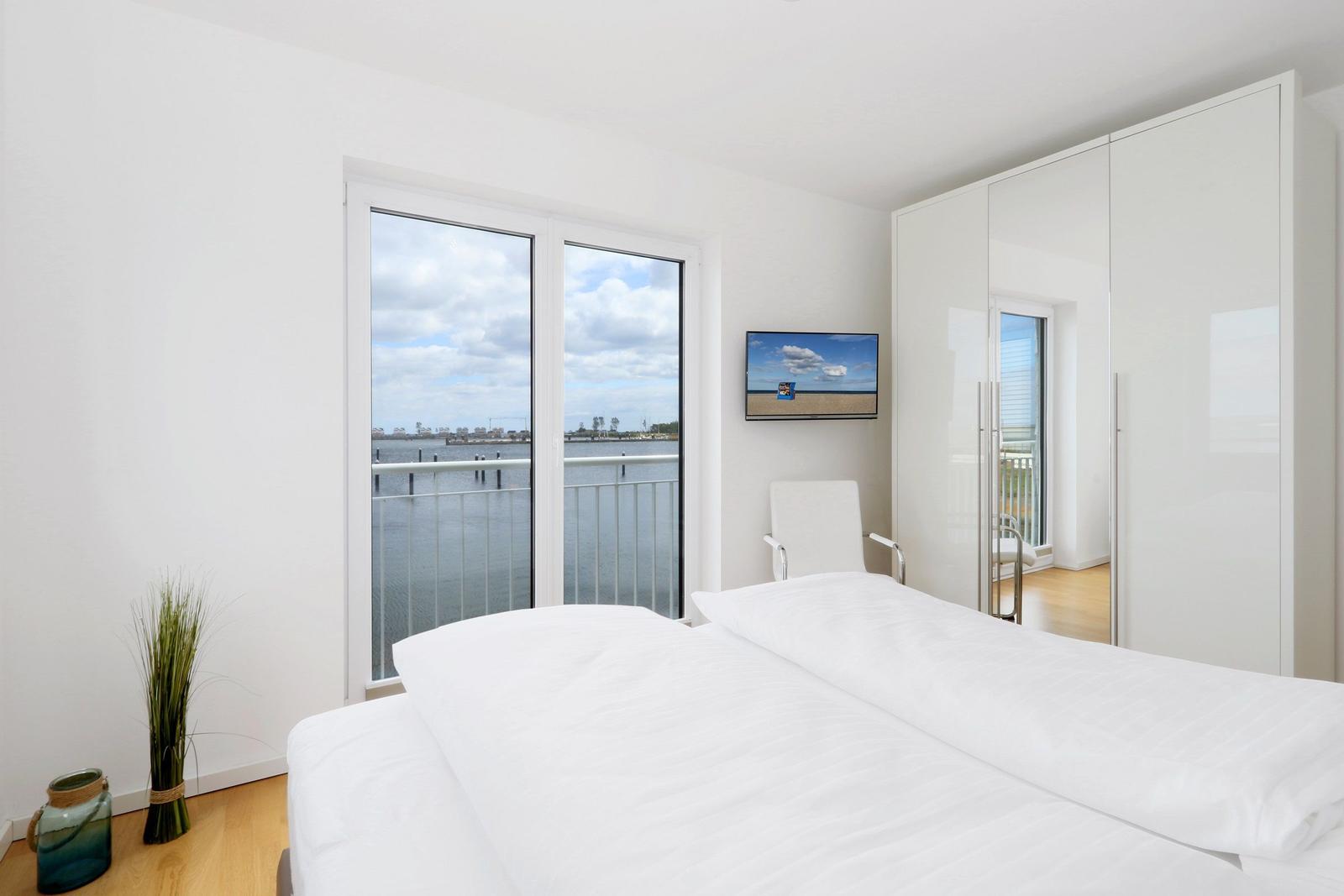 Schlafzimmer mit Blick auf Wasser, weißes Bett, Schrank und Fernseher an der Wand.