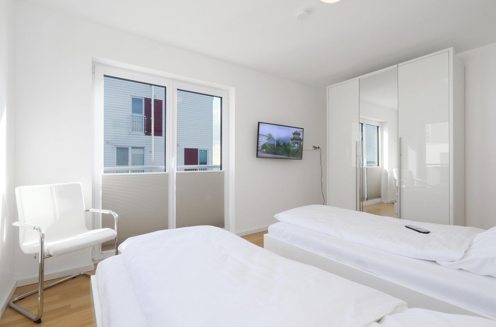 Zimmer mit zwei Betten, TV und Garderobe. Fenster mit Blick auf Gebäude.