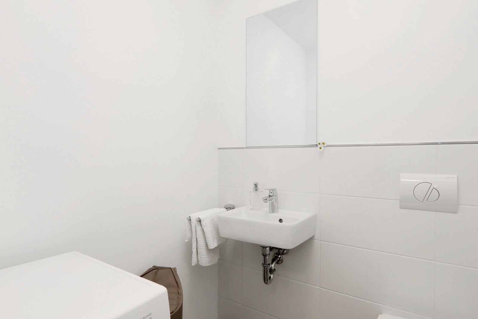 Badezimmer mit Waschbecken, Spiegel und Toilette. Weiße Fliesen und Handtuchhalter.