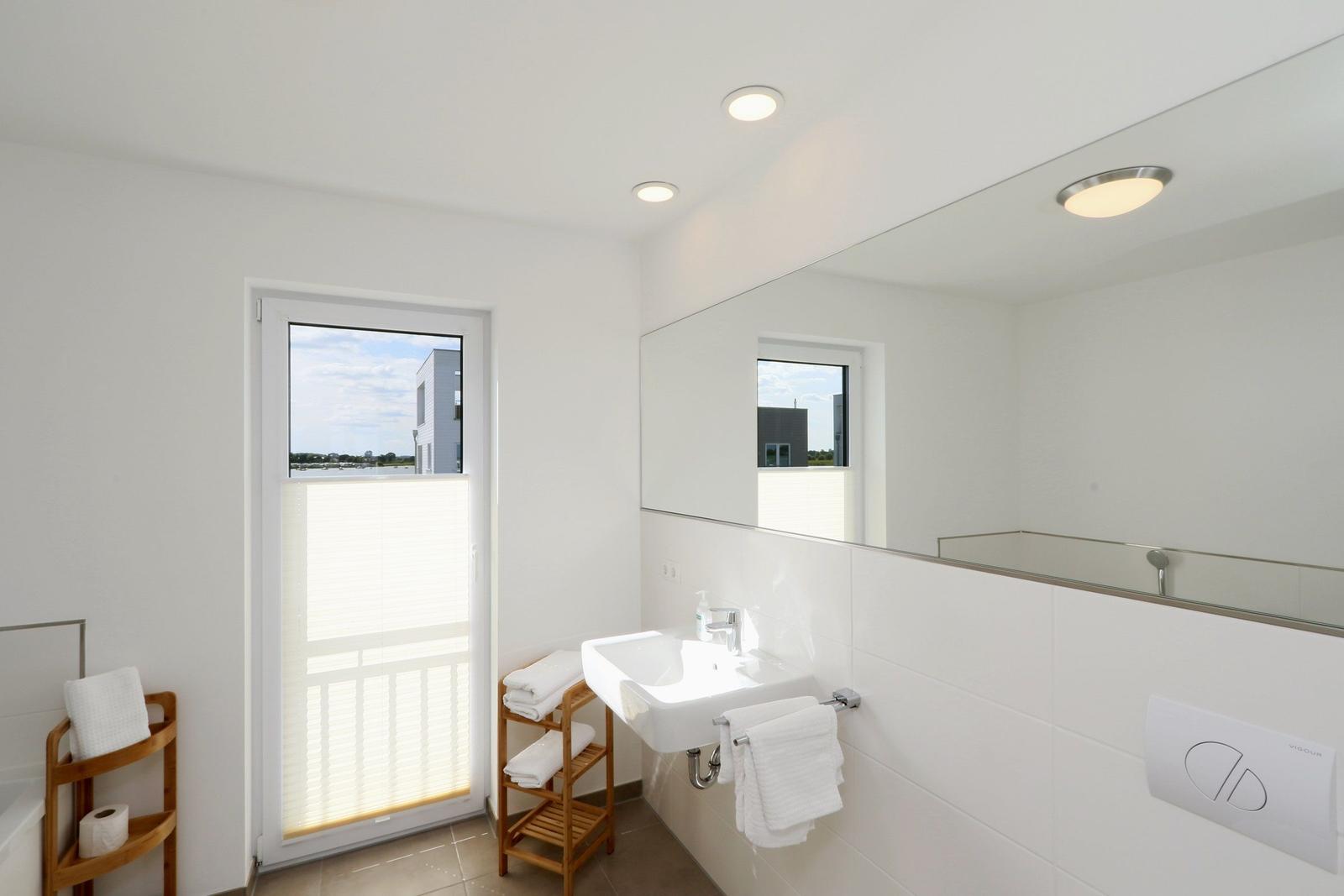 Badezimmer mit Waschbecken, Spiegel, Dusche und Fenster zum Balkon.