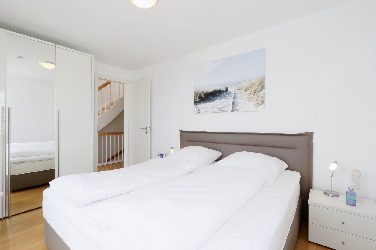 Schlafzimmer mit Doppelbett, Kleiderschrank und Wandbild. Tür führt zur Treppe.