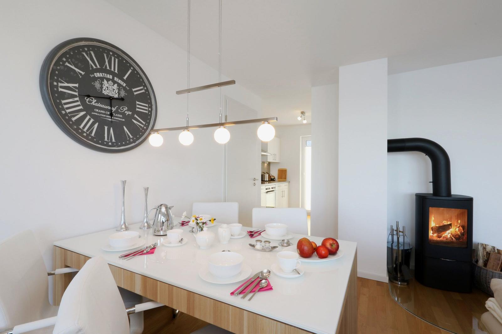 Esstisch mit weißen Stühlen, großer Wanduhr und Kaminofen im Hintergrund.