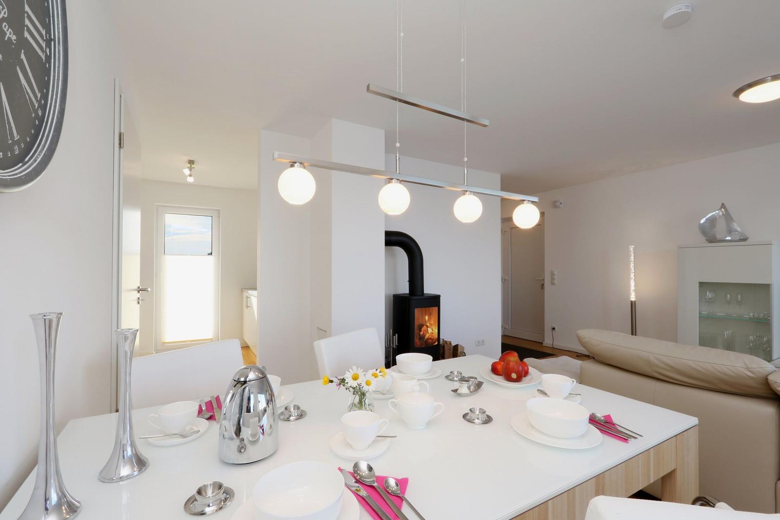 Essbereich mit Tisch, Stühlen und Kaminofen. Hintergrund zeigt Wohnbereich mit Sofa.