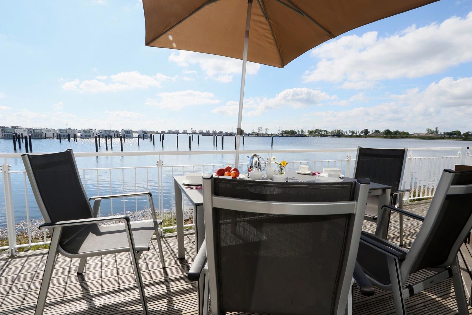 Terrasse mit Tisch, Stühlen und Sonnenschirm. Blick auf Wasser und gegenüberliegende Gebäude.