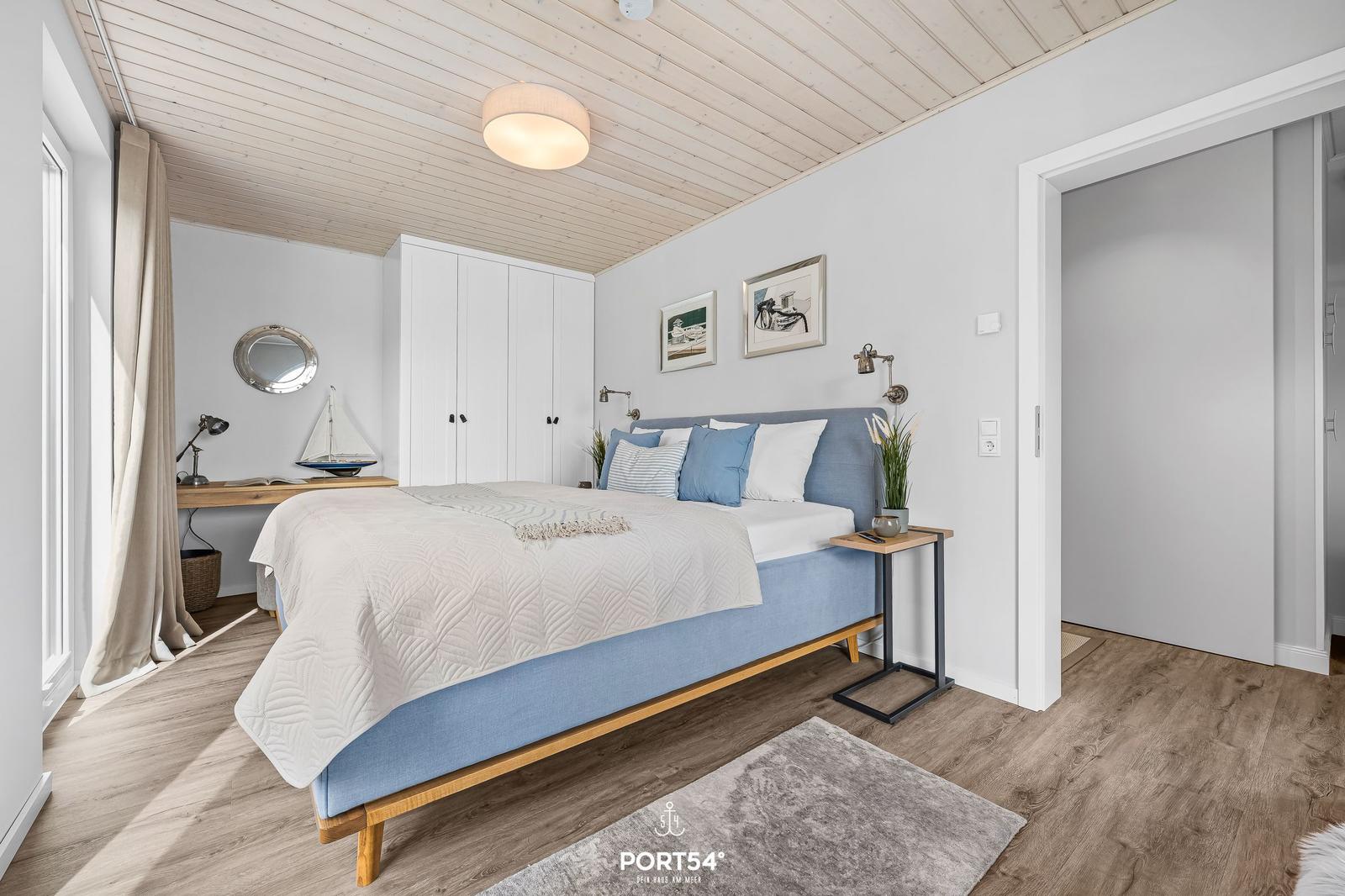 Schlafzimmer mit blauem Bett, Holzboden und weißen Wänden. An der Wand hängen zwei Bilder.