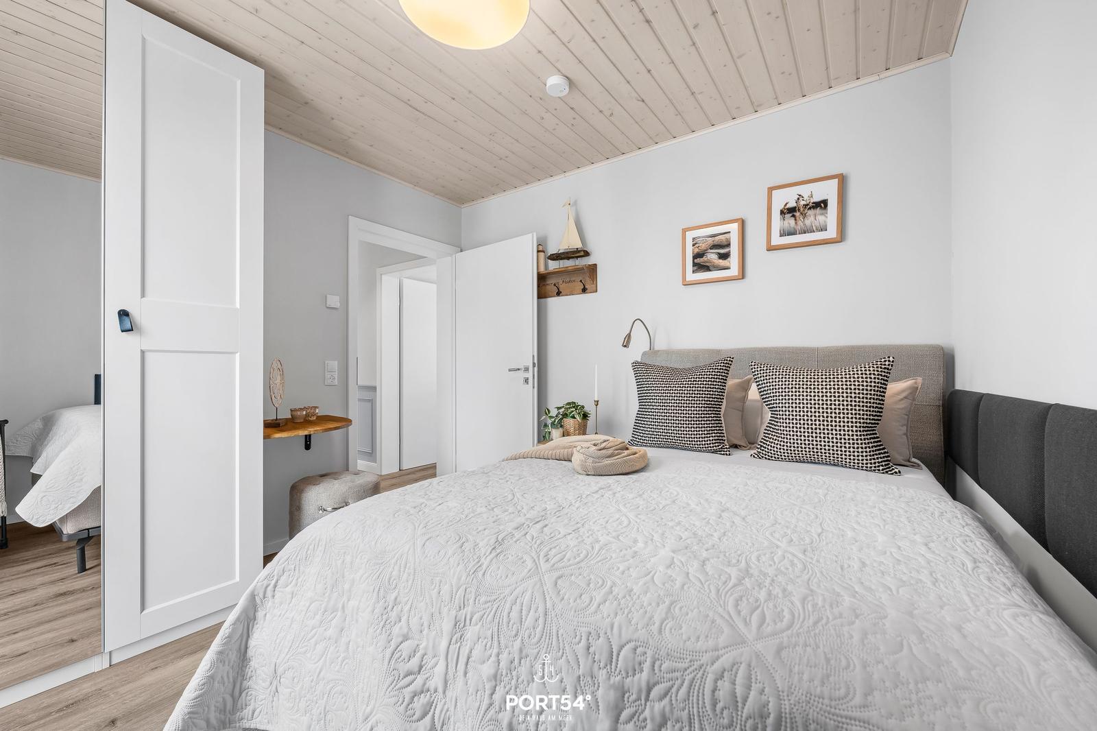 Schlafzimmer mit Bett, Wandbildern und Holzdecke. Tür führt in angrenzenden Raum.