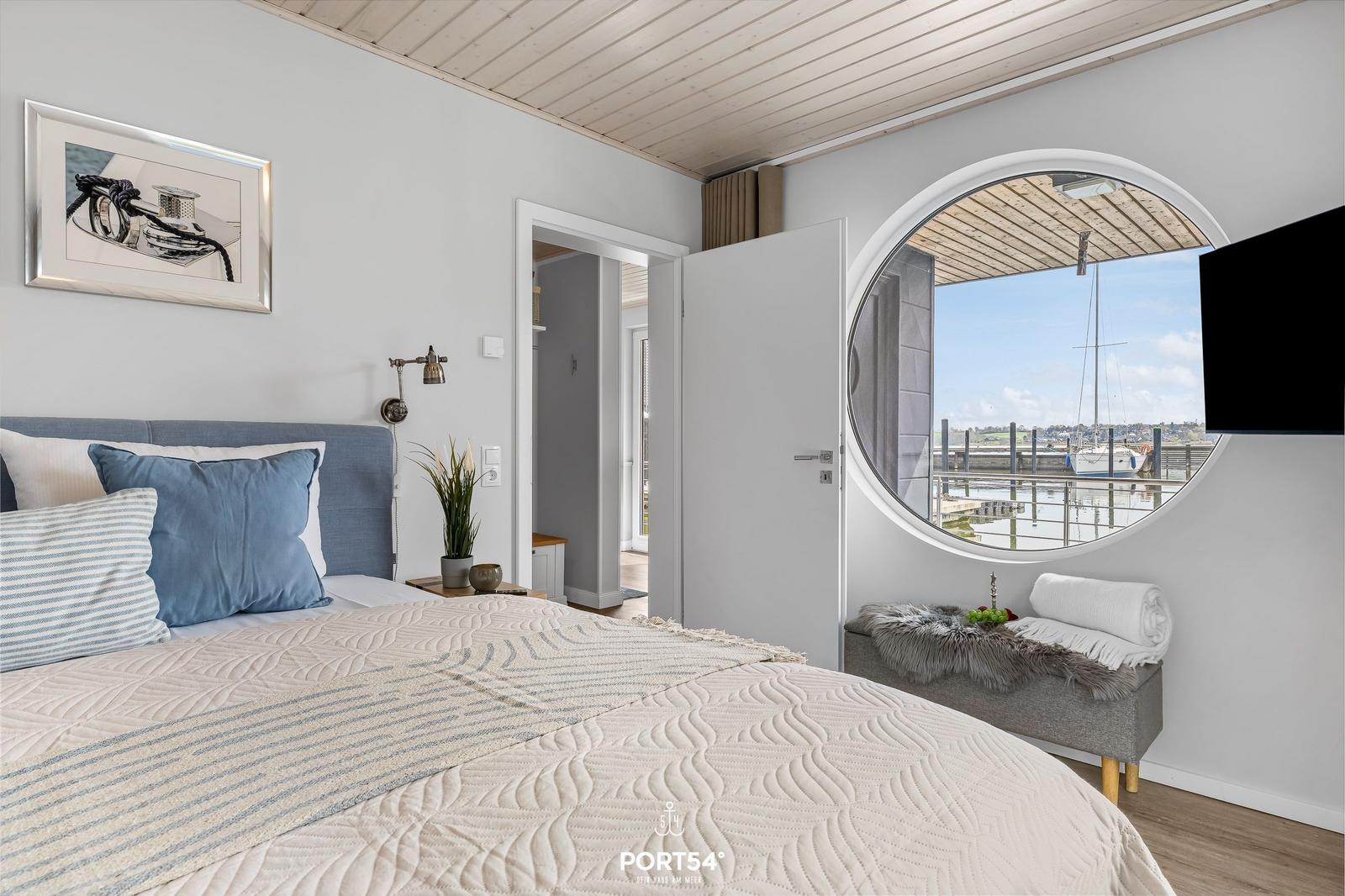 Schlafzimmer mit Rundfenster zum Hafen, Bett mit Kissen und Wandbild.