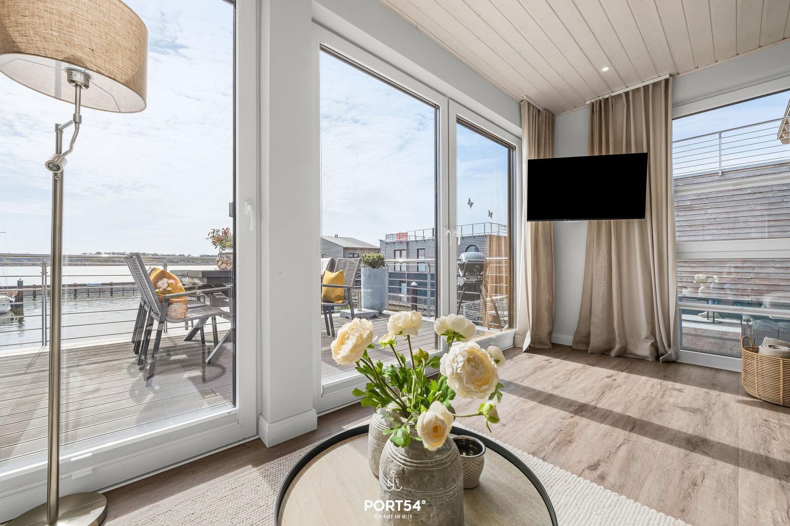 Wohnraum mit großem Fensterblick auf Terrasse und Wasser. TV, Tisch mit Blumen und Stehlampe.