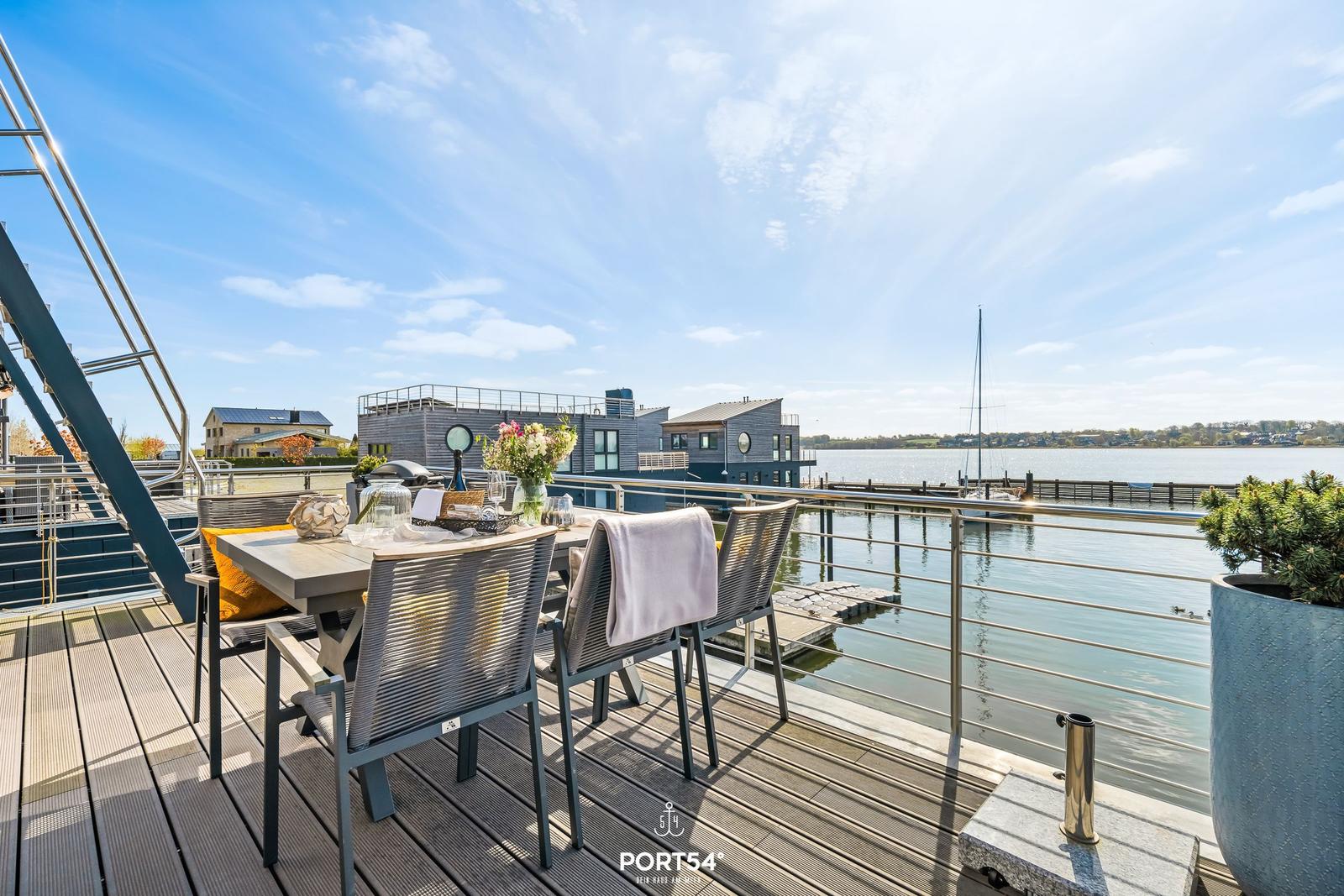 Terrasse mit Tisch und Stühlen am Wasser mit Blick auf Haus und Segelboot.