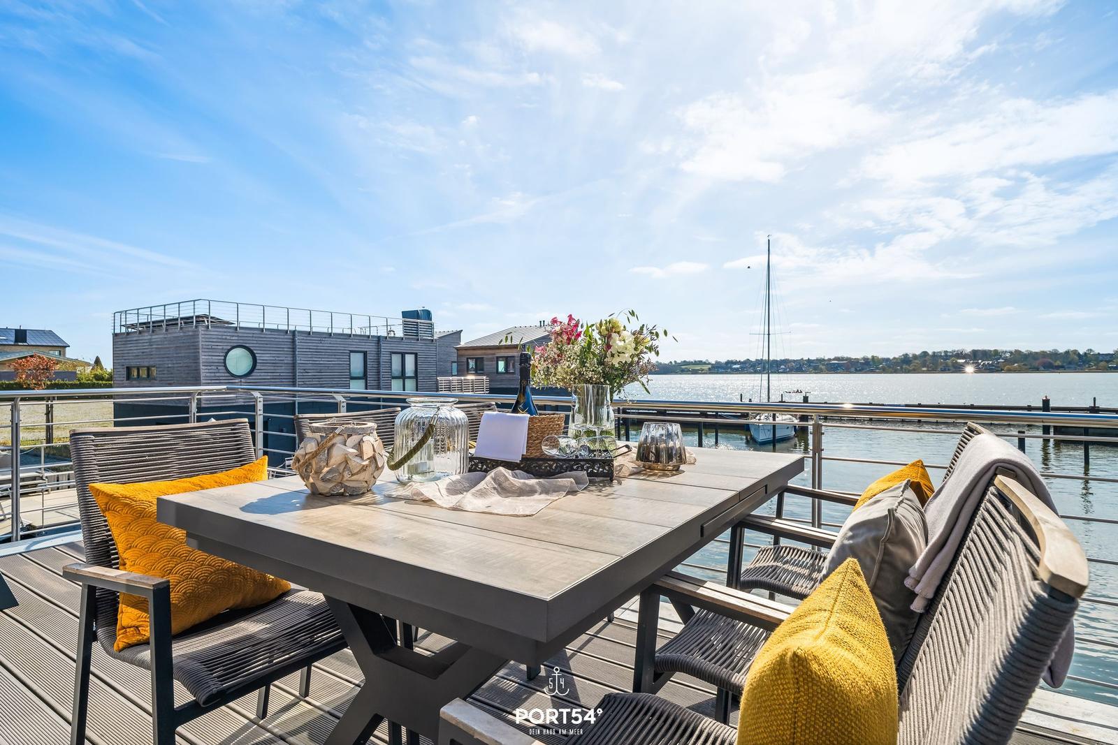 Terrasse mit Tisch und Stühlen, Blick auf Wasser und Boot.