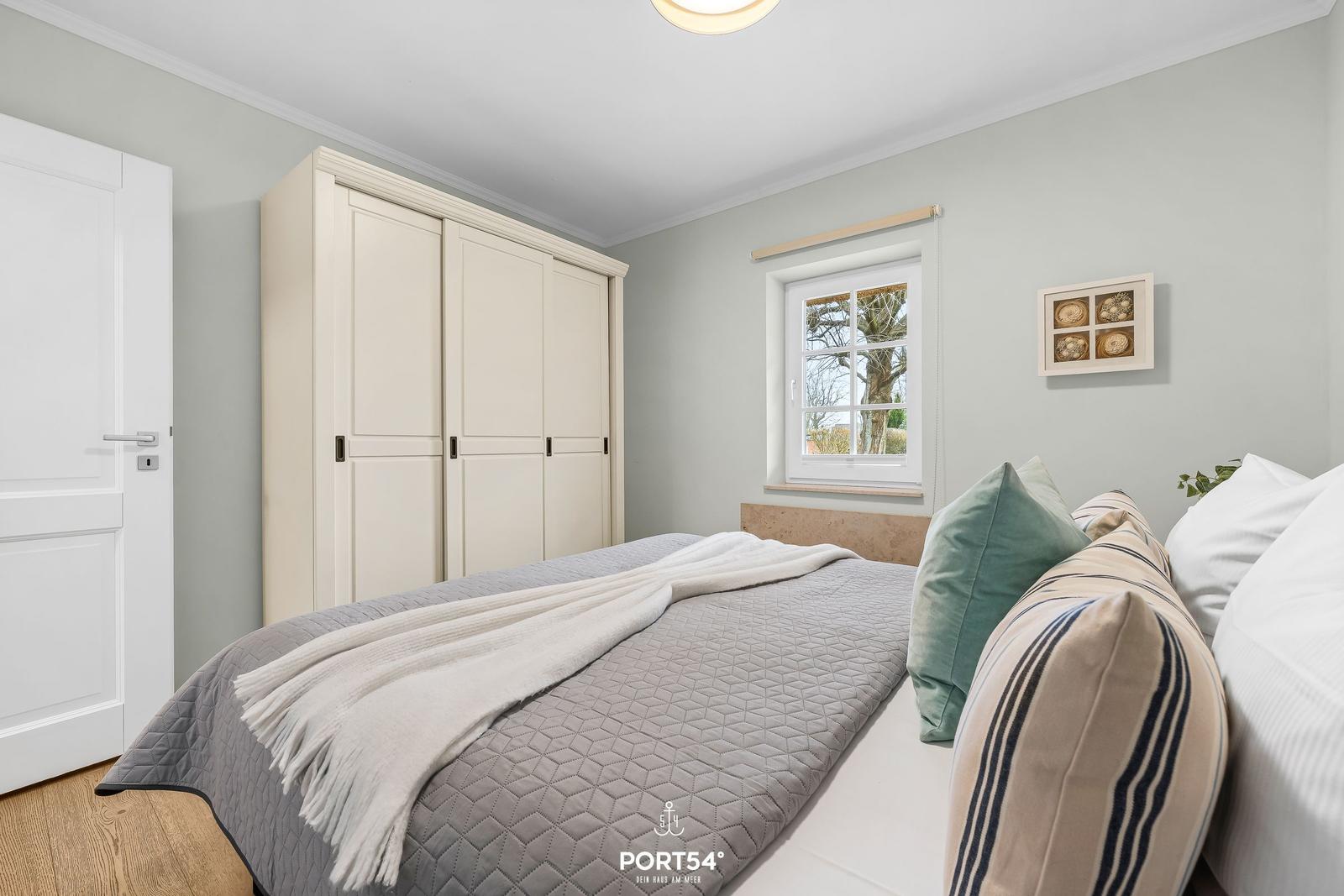Schlafzimmer mit Bett, Kleiderschrank und Fenster. Wandfarbe hellgrau.