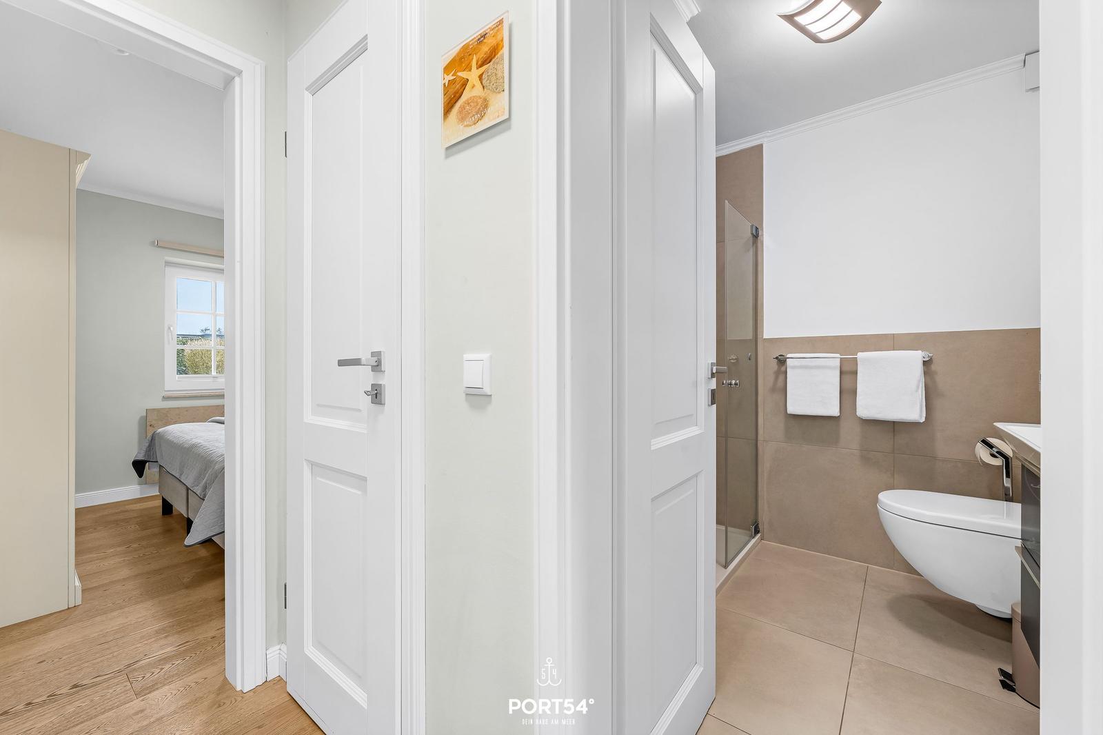 Badezimmer mit Dusche, Toilette und Handtuchständer. Durchgang zu Schlafzimmer mit Bett und Fenster.
