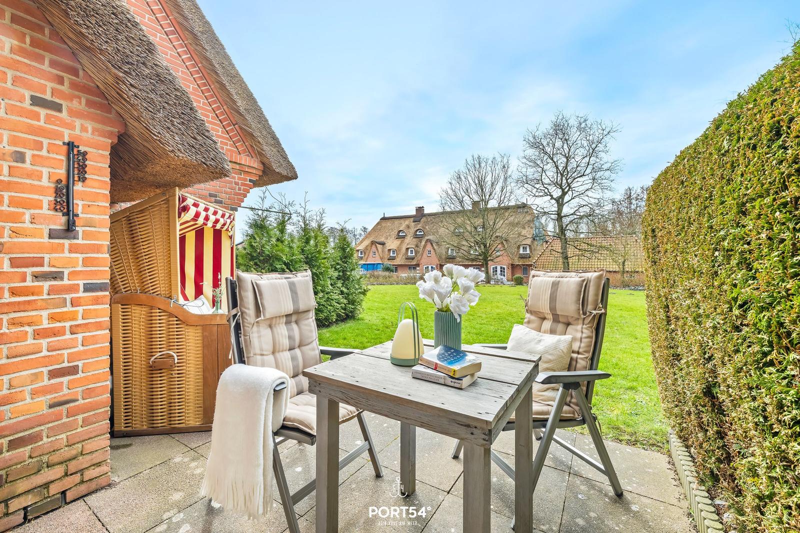 Terrasse mit Tisch, zwei Stühlen und Blick auf grünen Garten. Hintergrund: Haus mit Strohdach.