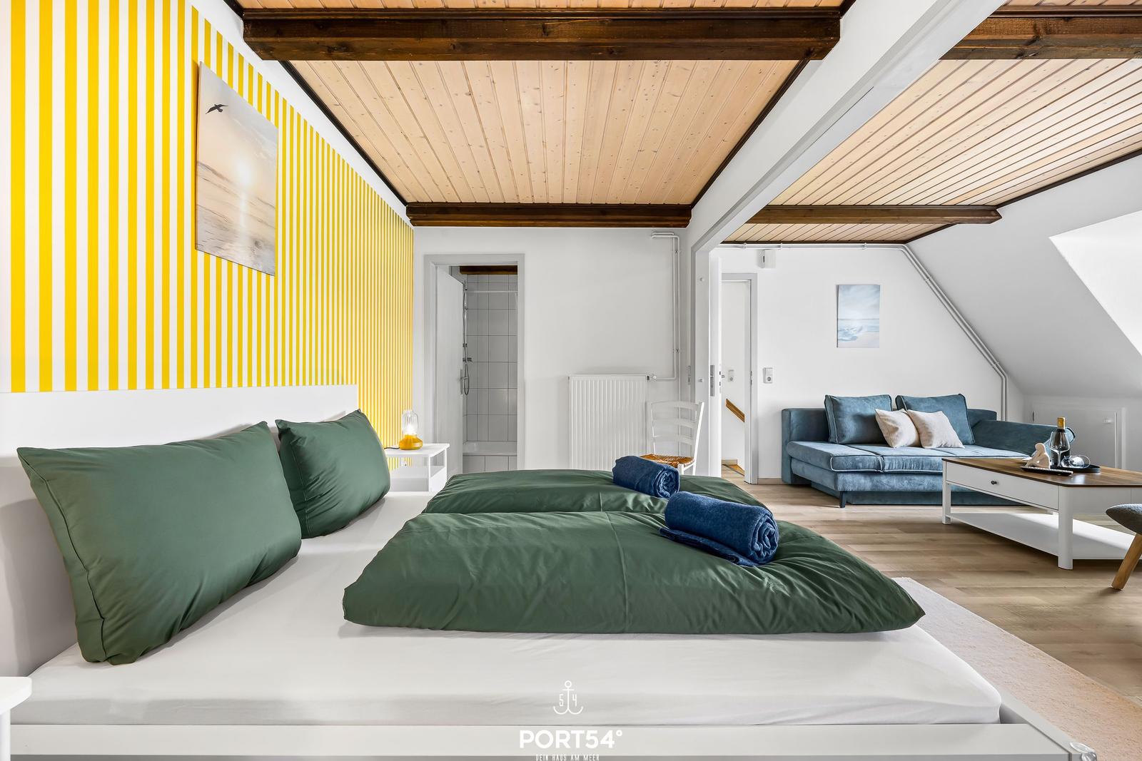 Schlafzimmer mit grüner Bettwäsche und gelb-weiß gestreifter Wand. Wohnbereich mit Sofa und Holzdecke.